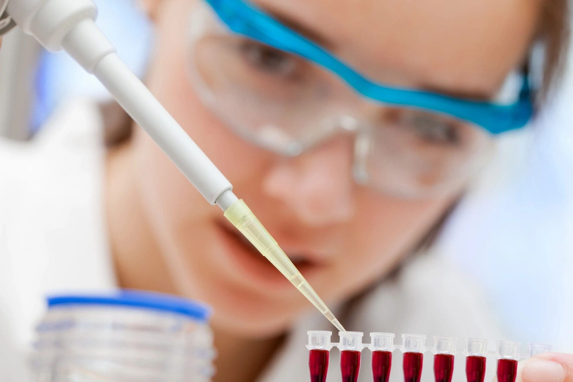 A woman in lab coat and goggles looking at test tubes.